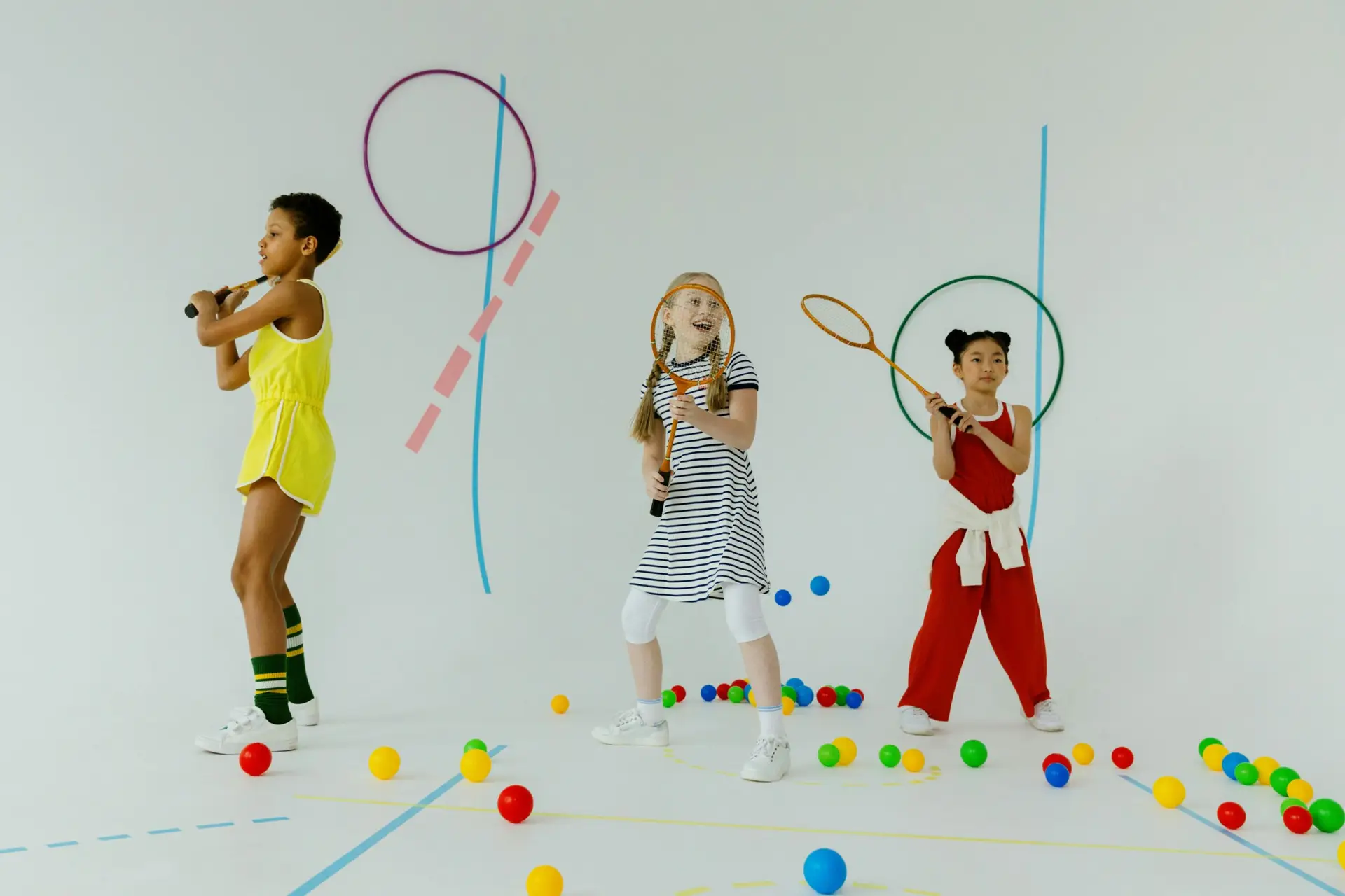 Three diverse children playing tennis indoors with rackets and colorful balls, showcasing fun and diversity.